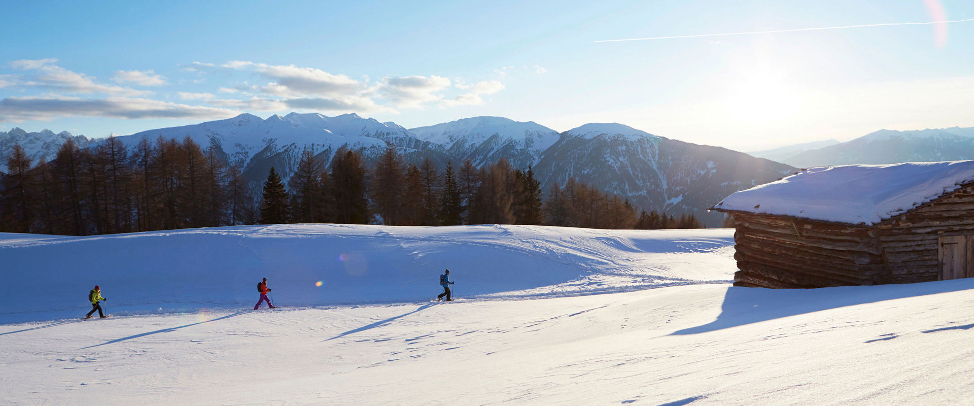 3 snowshoe hikers walk across a snow-covered meadow past a wooden hut at sunrise.