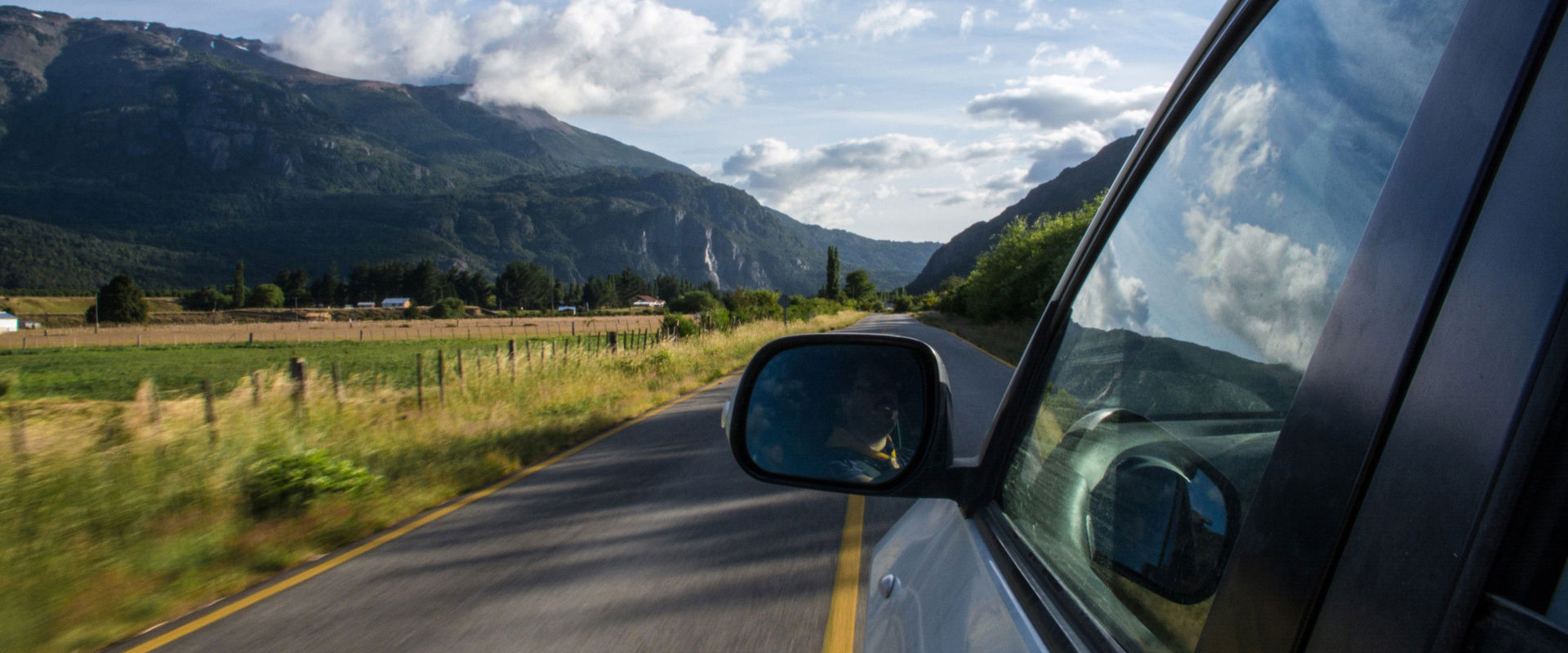 Car travelling on a straight. Photo from the left rear car window looking out onto the driver's side, the straight road and the surrounding area.
