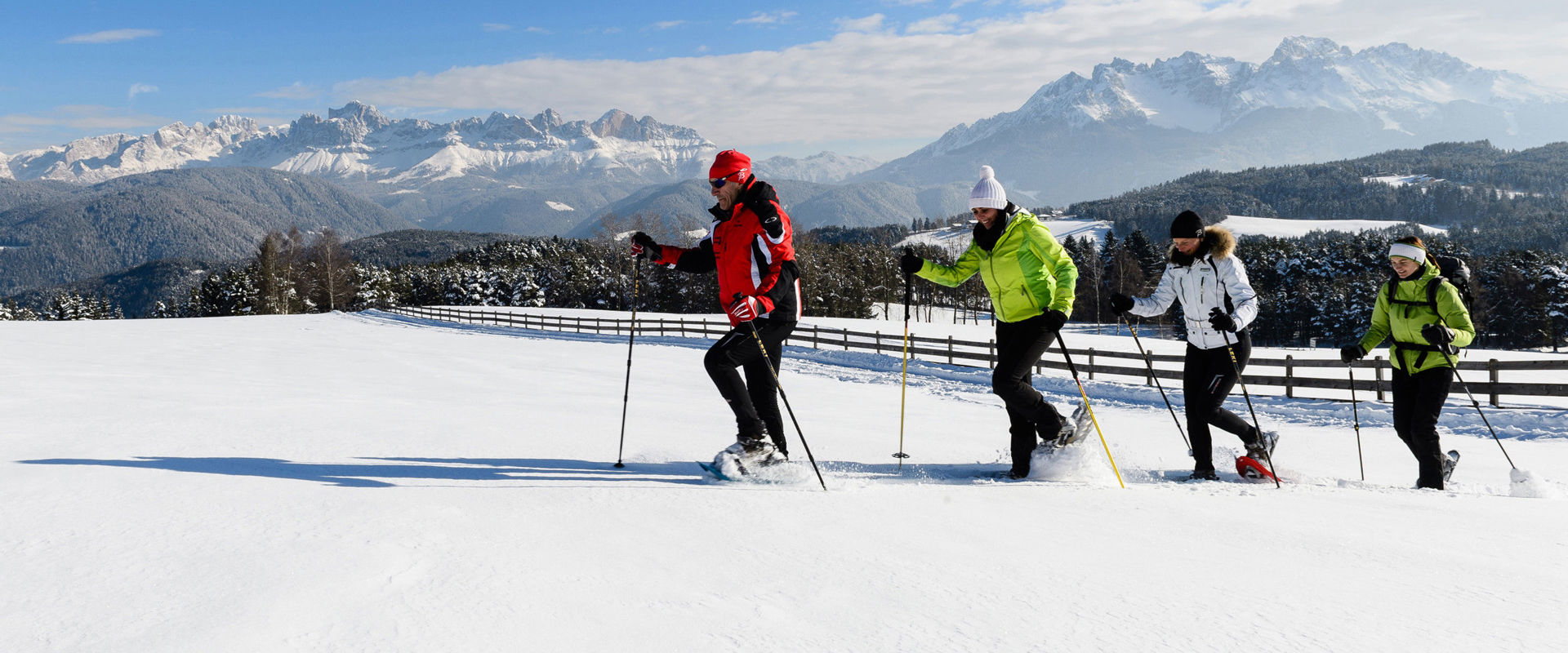 4 snowshoe hikers in deep snow on a fenced meadow. In the background, you can see a breathtaking mountain landscape.