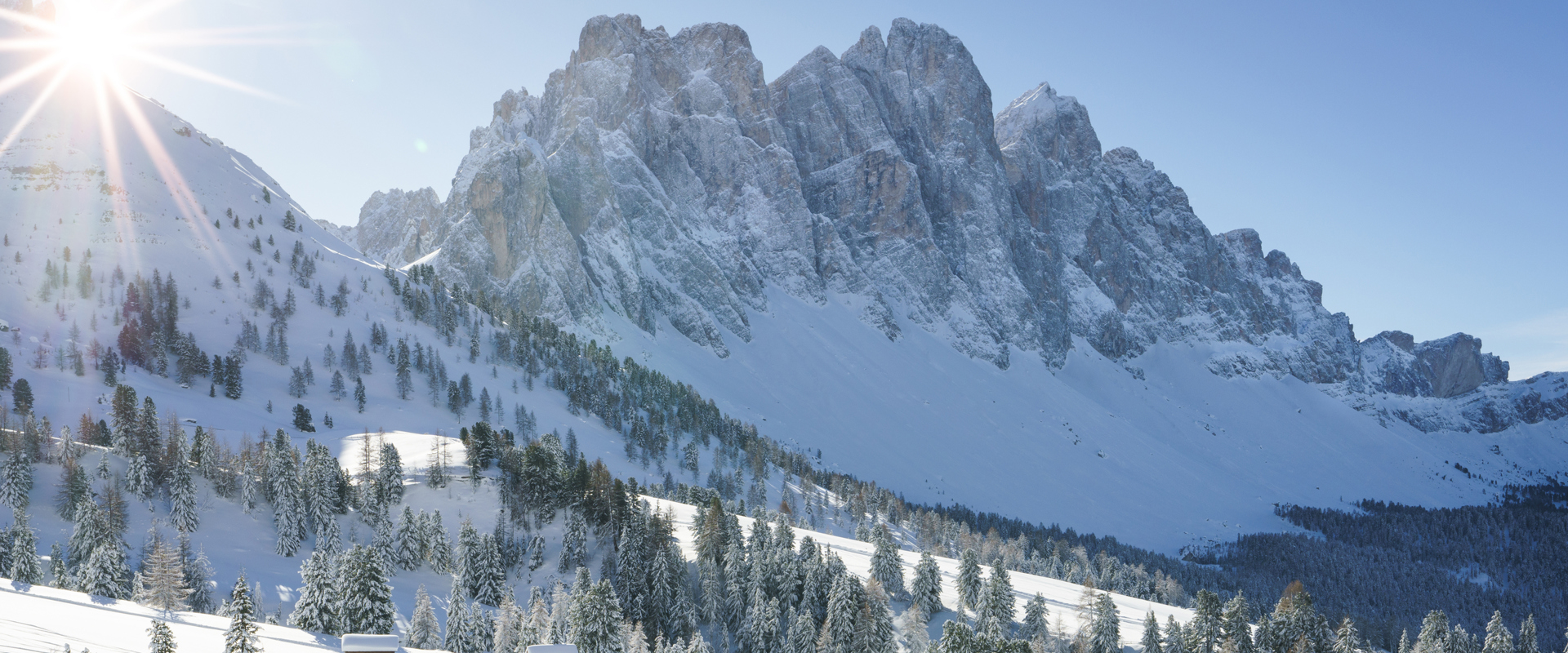 Villnöss Sunny, snow-covered mountain landscape with rocks and trees, with two ski tourers on the descent in front.
