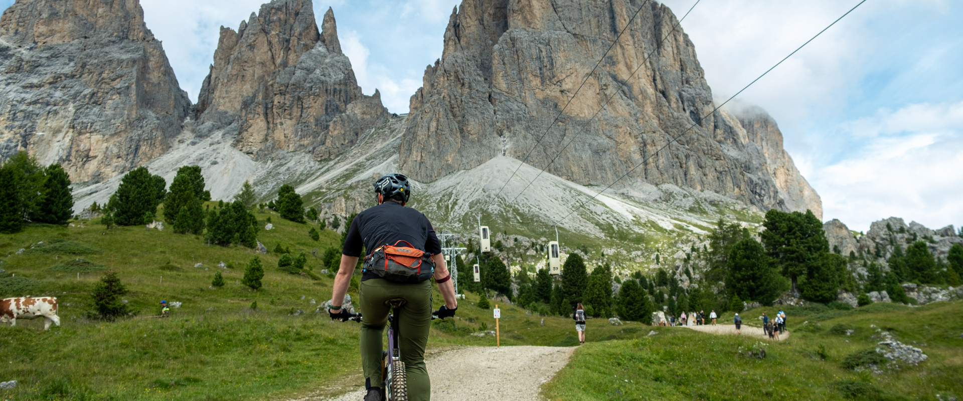 Mountain bikers on a path beneath a cable car and in front of the Dolomites